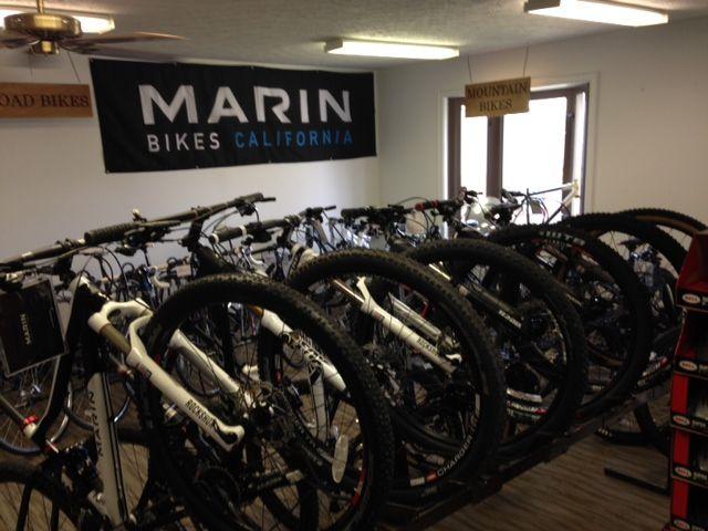 A display of various mountain bikes in a bike shop, with a prominent banner reading "MARIN BIKES CALIFORNIA." The bikes are arranged in rows, showcasing their features, with some visible brand labels and tires. Signs indicating "Road Bikes" and "Mountain Bikes" are also present in the background.