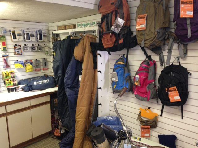 A cluttered outdoor retail store interior featuring various backpacks of different colors hanging on the wall, sleeping bags displayed on a rack, and camping gear on shelves. The store has a neatly arranged counter in the foreground, showcasing additional camping equipment and supplies.