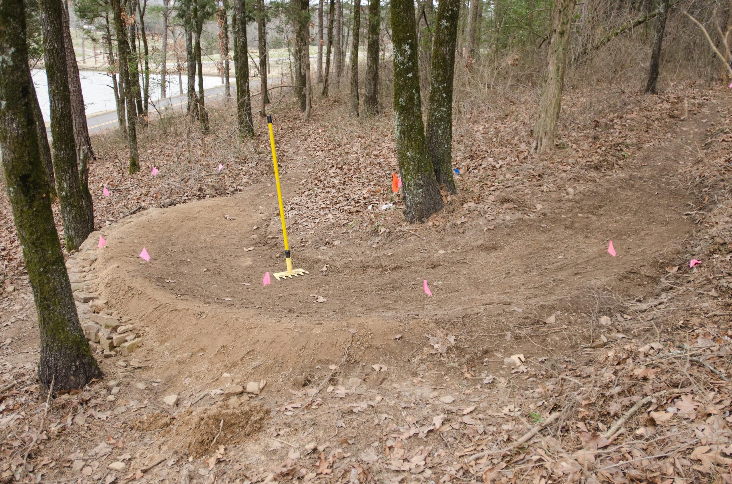 A dirt path curves through a forested area, marked by small pink flags along its edges. A yellow landscaping rake stands upright on the ground. The scene includes trees with scattered leaves on the ground, indicating a recent clearing or construction activity in this natural setting. A road can be seen in the background. Old Post mountain bike trail.