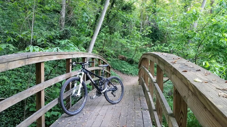 A black mountain bike leaning against a wooden bridge in a lush green forest. The bridge arches over a path surrounded by trees and dense vegetation, creating a peaceful, natural setting. Buttermilk mountain bike trail.