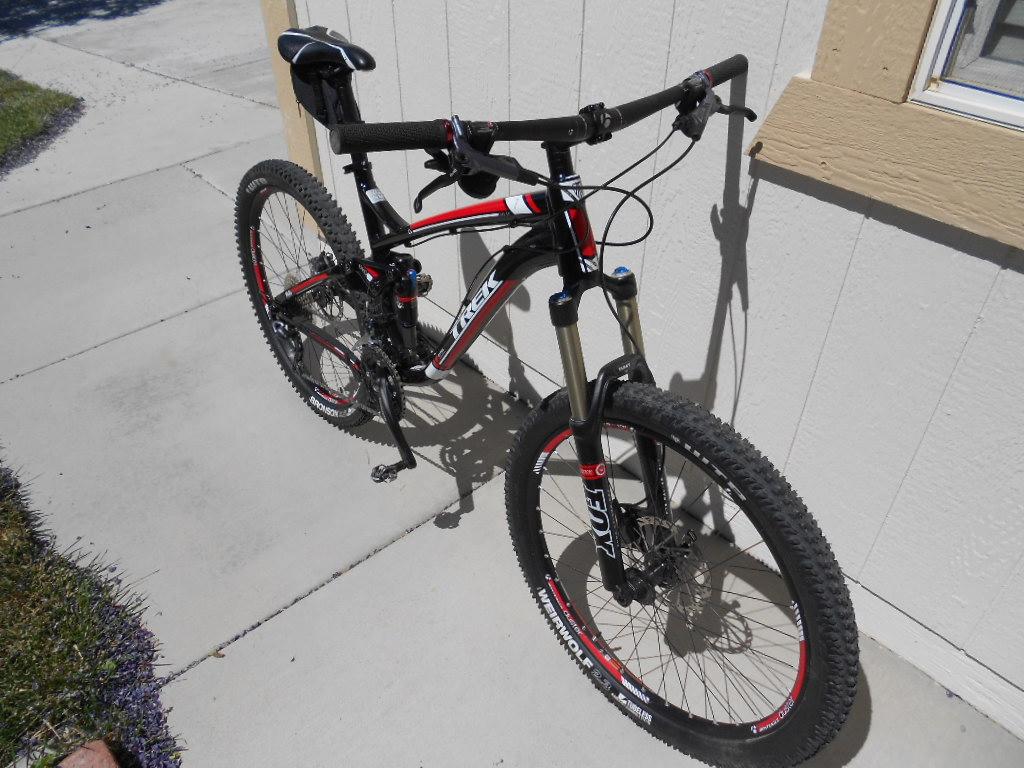 Trek Fuel EX 7: A black and red mountain bike is parked on a concrete surface next to a light-colored wall. The bike features thick tires and a suspension fork, designed for off-road cycling. Sunlight casts a shadow of the bike on the ground, and greenery is visible in the background.