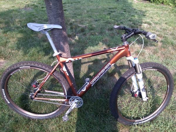 Gary Fisher Rig 29er: A brown mountain bike parked on grass near a tree. The bike features a lightweight frame, thick tires, and front suspension, showcasing its design for off-road trails and rugged terrain.