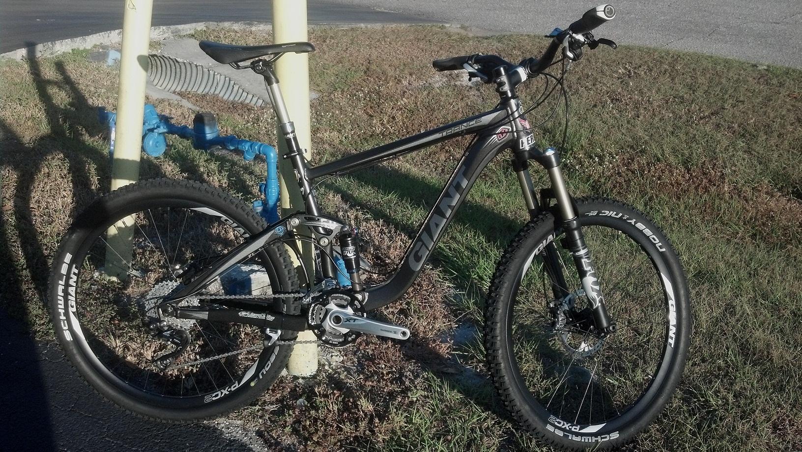 Giant Trance X1: A black mountain bike with suspension and large tires is parked beside a yellow pole and blue water pipes in a grassy area.