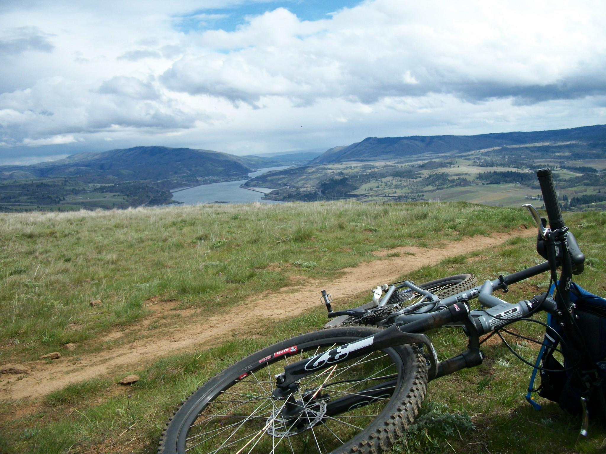 A scenic view from a hilltop overlooking a river, with sprawling green fields and mountains in the background. In the foreground, two mountain bikes are resting on the grass, positioned near a dirt trail. The sky is partially cloudy, creating a serene outdoor atmosphere. Syncline mountain bike trail.