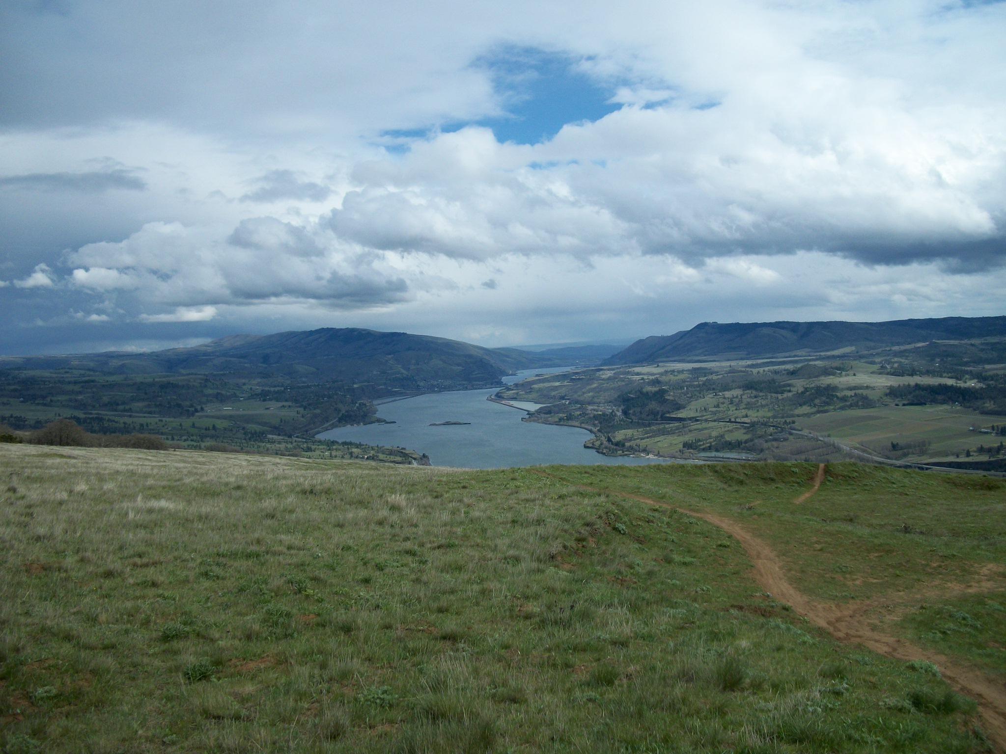 A panoramic view of a river winding through lush green valleys, with rolling hills and mountains in the background under a partly cloudy sky. A dirt path leads down the grassy slope in the foreground. Syncline mountain bike trail.