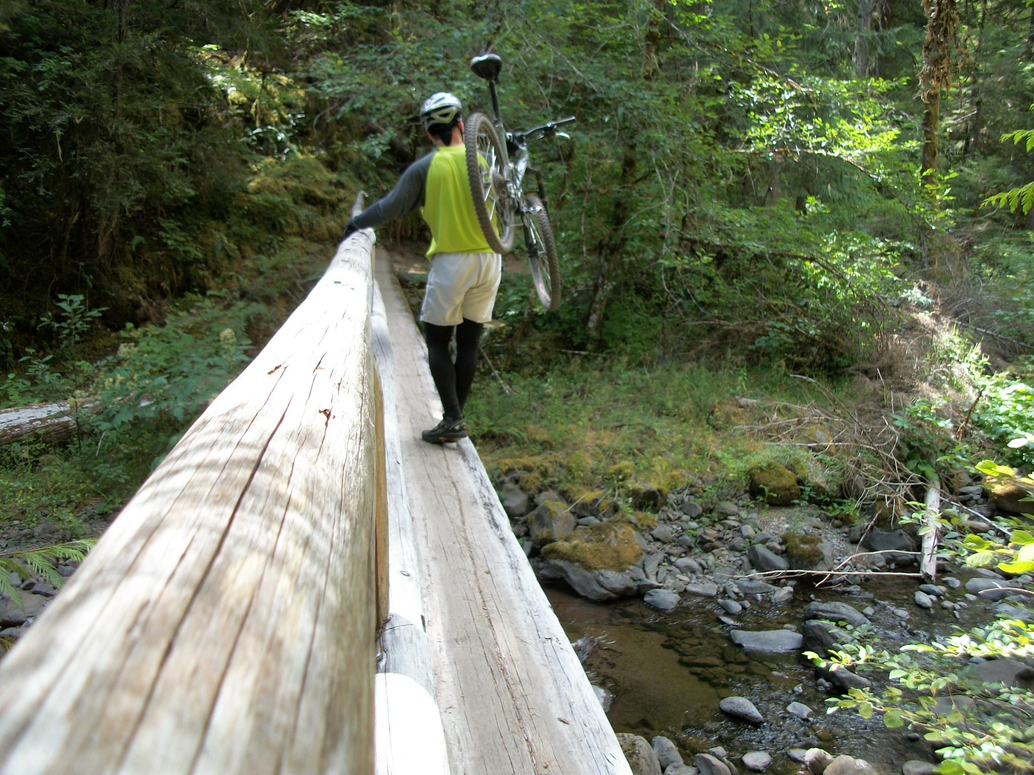 A person walking on a log bridge in a lush forest, carrying a bicycle. The scene features dense greenery, with sunlight filtering through the trees, creating a serene outdoor atmosphere. The individual is wearing a helmet, a bright vest, and athletic shorts, suggesting an active outdoor adventure. Middle Fork Trail mountain bike trail.