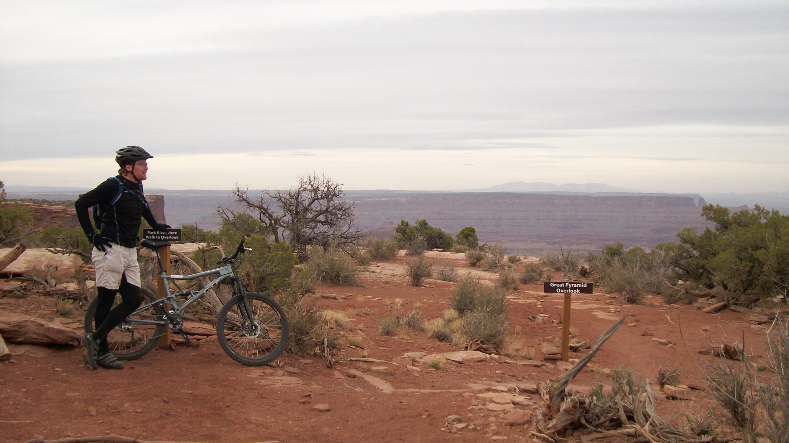 A mountain biker pauses at a scenic overlook, standing next to his bike on a rocky, dry terrain. A sign indicates the bike parking area and the path to the Great Pyramid Overlook in the background, showcasing a vast landscape with mesas under a cloudy sky. Vegetation includes sparse shrubs and trees typical of arid environments. Dead Horse Point State Park mountain bike trail.