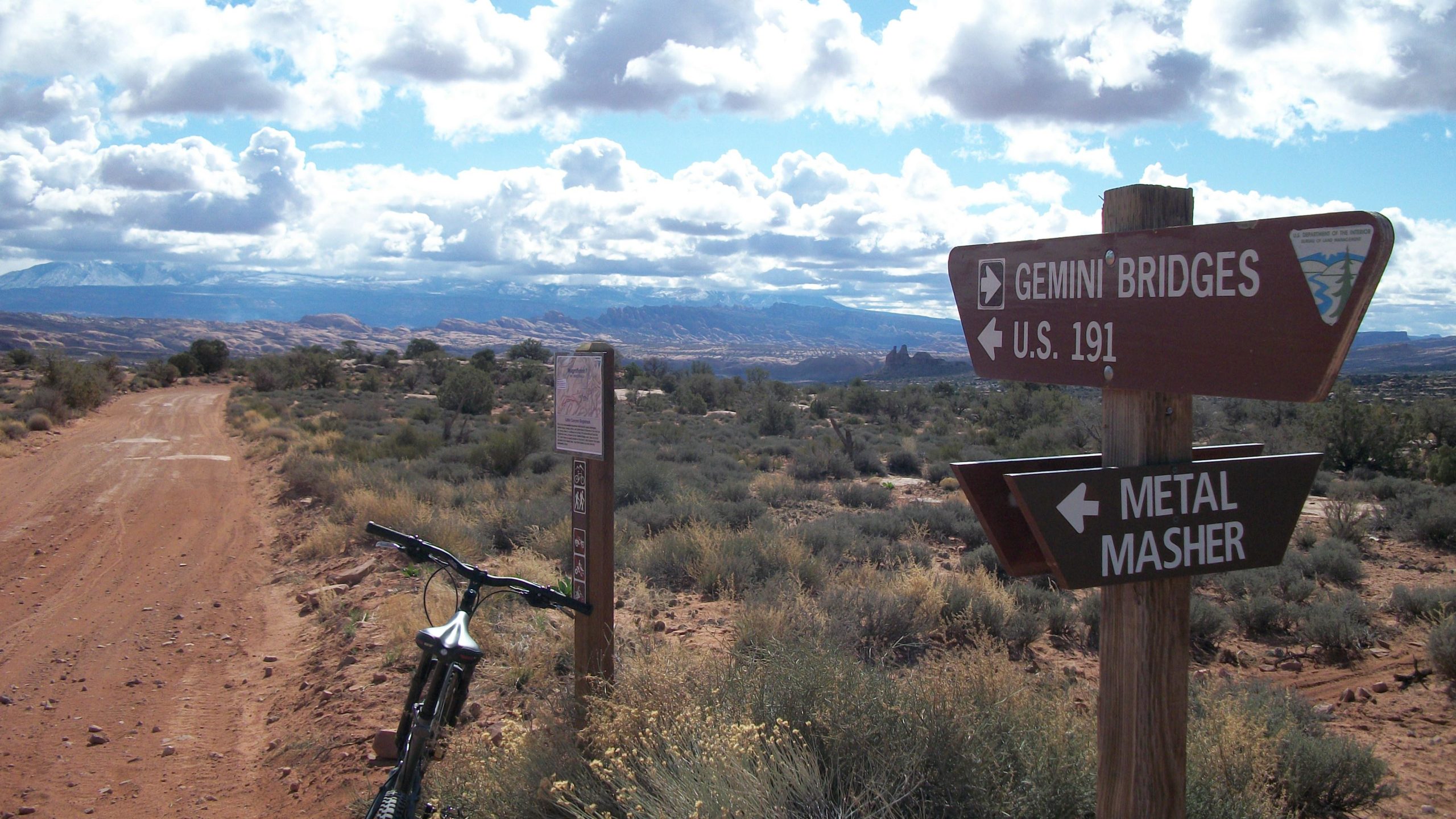 A dirt trail leading through a desert landscape, with two directional signs. One sign points to Gemini Bridges and U.S. 191, while the other points to Metal Masher. A mountain bike rests against the sign, surrounded by sparse vegetation and a backdrop of rocky hills and clouds in the sky. Gemini Bridges mountain bike trail.