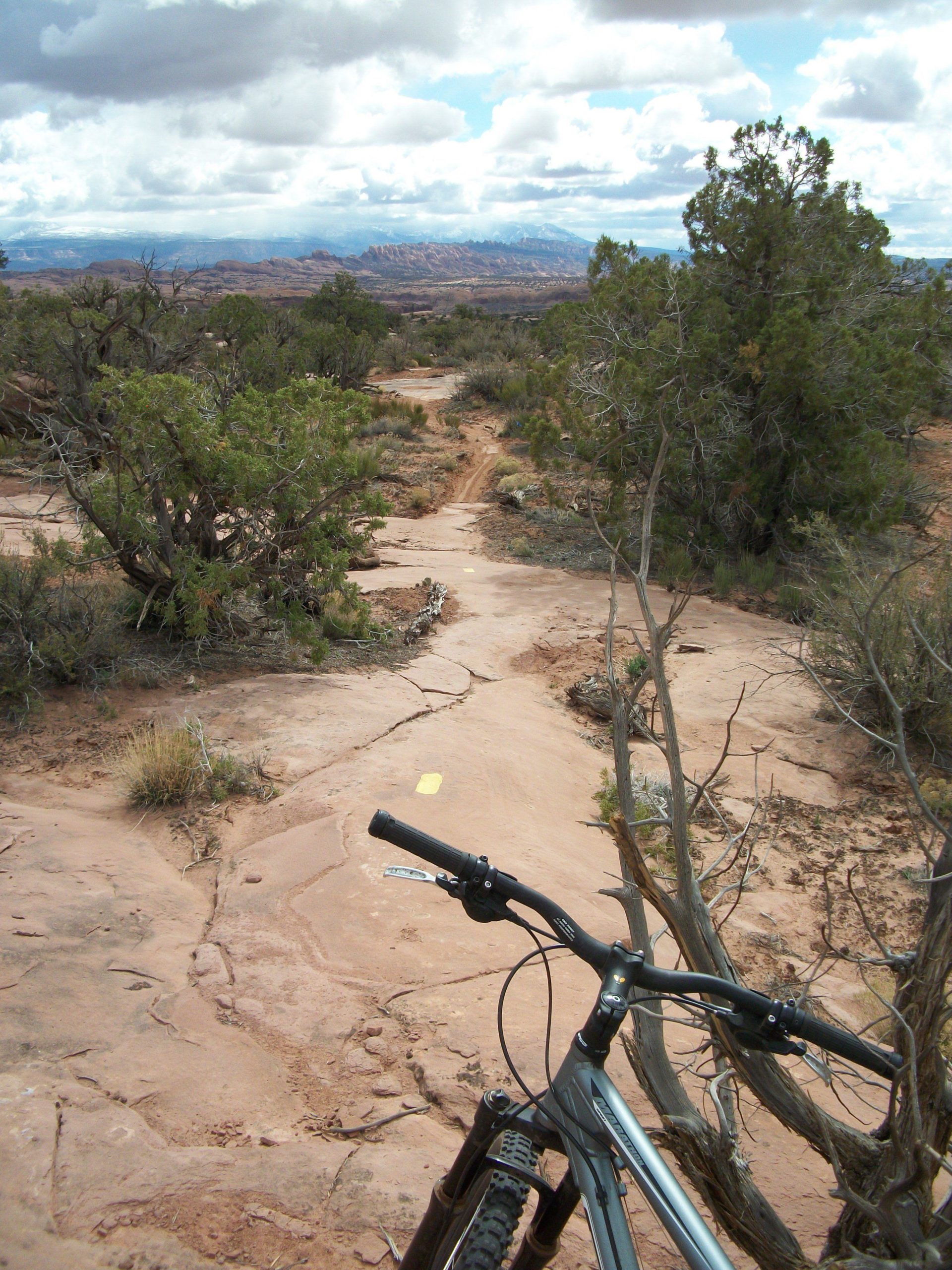 A mountain bike positioned on a rocky trail surrounded by desert vegetation, with a distant view of layered hills and mountains under a cloudy sky. A yellow trail marker is visible on the ground, guiding the path. Gemini Bridges mountain bike trail.
