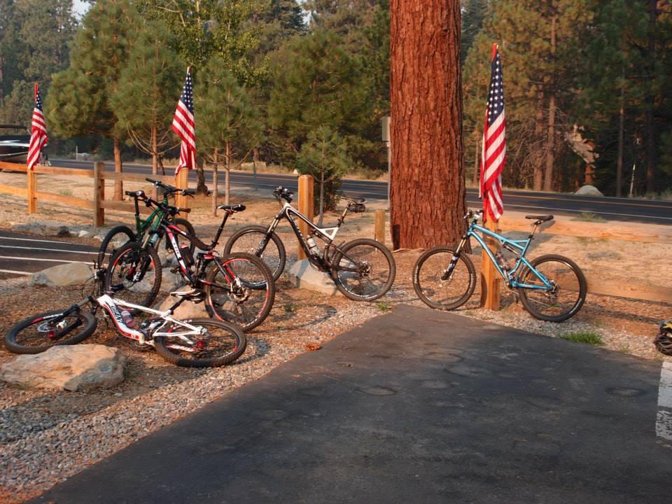 A collection of mountain bikes parked on a gravel patch with a backdrop of trees and American flags. The scene captures a peaceful outdoor setting, indicating a popular biking area. Tahoe Rim Trail: Tahoe Meadows to Tunnel Creek Road / Flume Trail mountain bike trail.