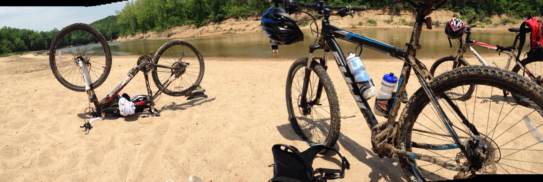 Fuji nevada 1.5 29er: A panoramic view of two mountain bikes parked on sandy shores near a river. One bike is lying on its side with the front wheel up, showcasing mud on its frame, while the other bike stands upright. In the background, a lush green forest borders the riverbank, and a person can be seen wading in the water.