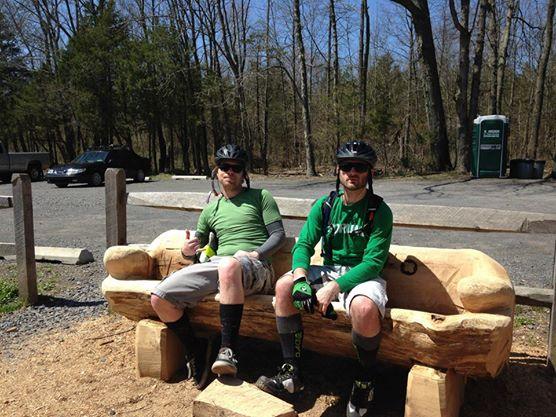 Two men wearing cycling helmets and outdoor clothing sit on a wooden log bench surrounded by trees. They are posing with relaxed expressions, one giving a thumbs-up. A parking area is visible in the background, along with a portable restroom. It’s a sunny day, suggesting they're taking a break from biking or outdoor activities. Nockamixon State Park mountain bike trail.
