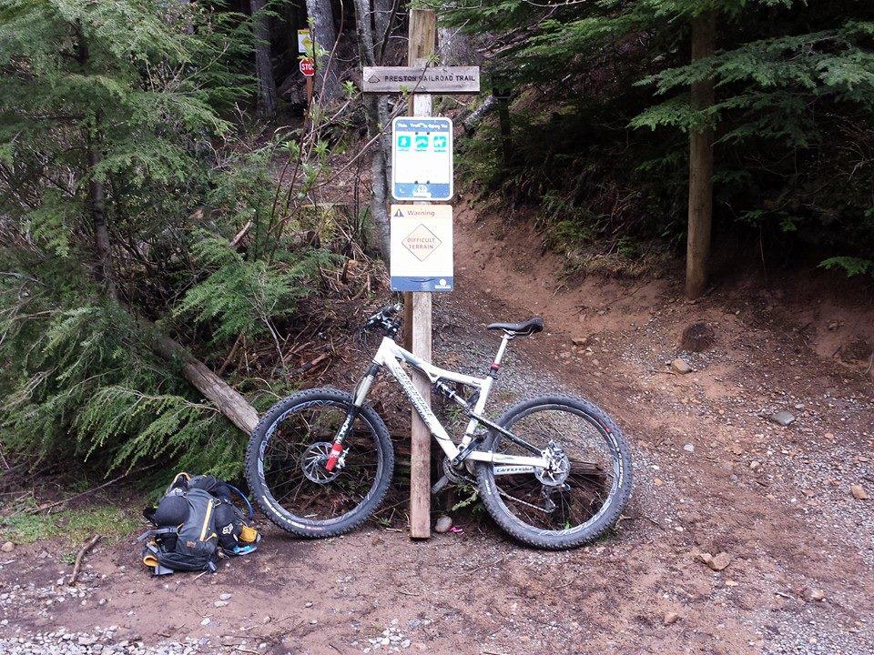 Cannondale Rize 3: A mountain bike leaning against a trail sign in a wooded area. The sign indicates the entrance to Preston Trail and includes warnings about difficult terrain. A black backpack is placed on the ground nearby, and the surroundings are lush with trees and gravel.