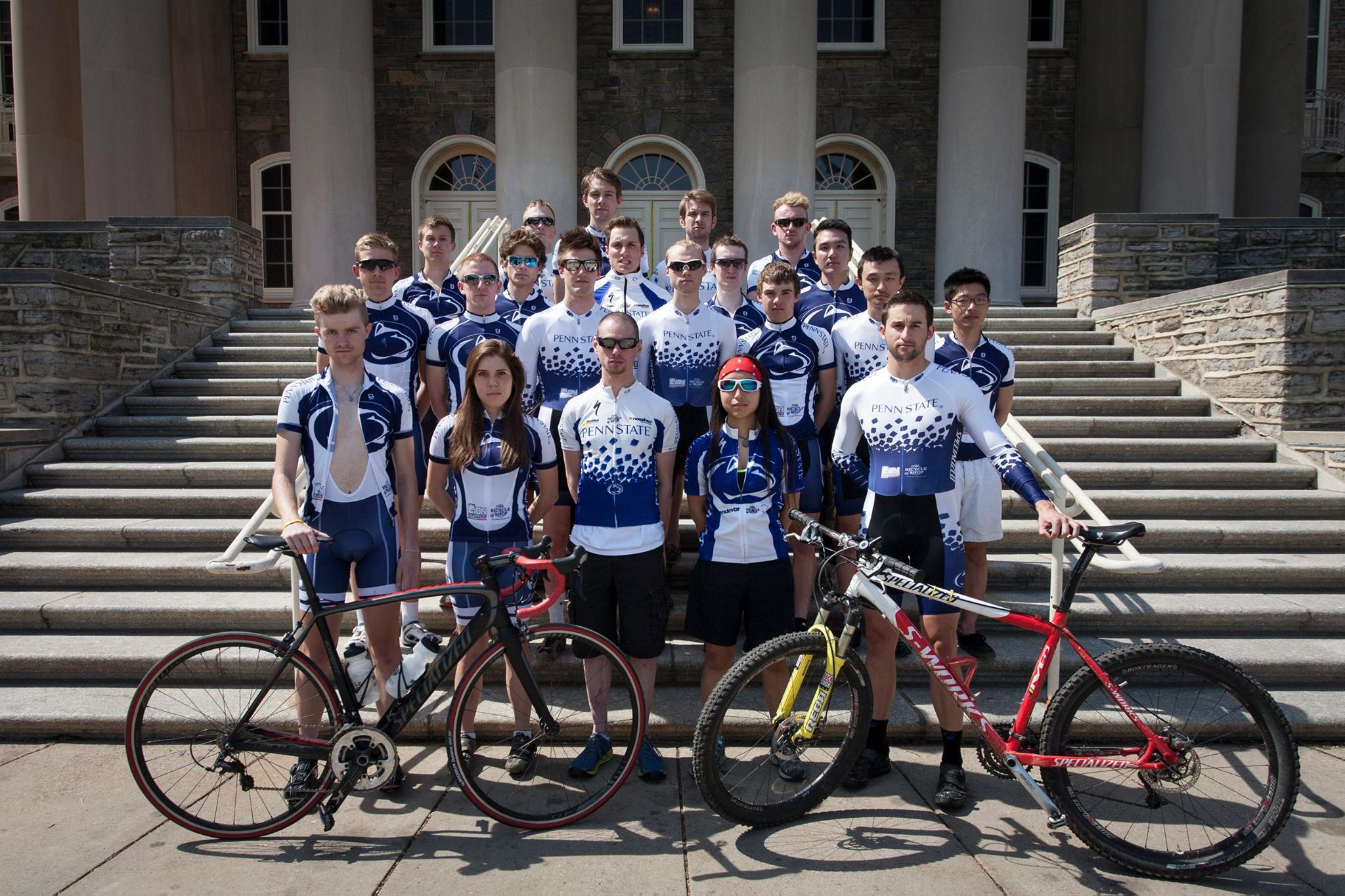 Group photo of a cycling team wearing matching blue and white jerseys with the "Penn State" logo, standing in front of a stone building with large columns. The team members pose on steps, holding bicycles, with some members in the forefront displaying road and mountain bikes.
