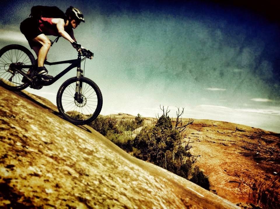 A mountain biker rides down a rocky slope against a backdrop of blue sky and rugged terrain, showcasing the intensity and thrill of outdoor biking. Slickrock mountain bike trail.