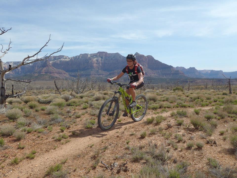 A person riding a mountain bike on a dirt trail in a desert landscape, with sparse vegetation and dry terrain. The background features striking red rock formations and distant mountains under a clear blue sky.