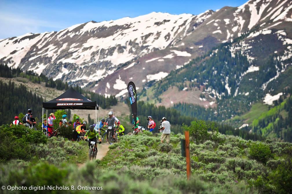 A scenic mountain landscape featuring a group of mountain bikers and spectators gathered near a tent with the "Maxxis" logo. Snow-capped mountains rise in the background, while vibrant green hills and shrubs frame the scene. Several cyclists are riding along a dirt trail, showcasing an active outdoor event. Rim Trail mountain bike trail.