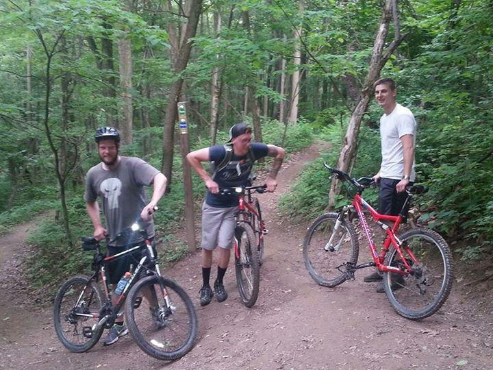 Three young men stand with their mountain bikes on a dirt trail in a lush green forest. They appear to be resting or chatting, with trees surrounding them and a trail marker visible in the background. The scene captures a moment of camaraderie in an outdoor setting. Allegrippis Trails mountain bike trail.
