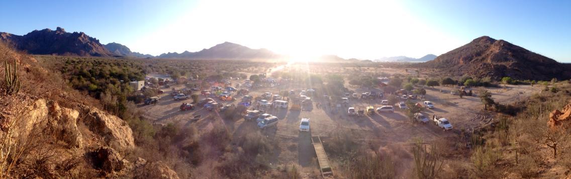 A panoramic view of a desert campsite during sunset, featuring several parked vehicles amidst rugged terrain and mountainous backgrounds. The sun is shining brightly, casting warm light over the campsite, with cacti and sparse vegetation visible. Rancho Nacapule - 12 Horas Salvando Vidas 2014 mountain bike trail.