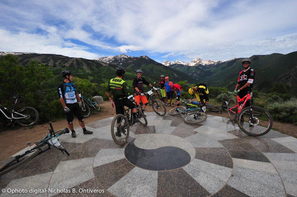 A group of mountain bikers gathered at a scenic viewpoint surrounded by lush green hills and snow-capped mountains in the background. The cyclists, wearing colorful jerseys and helmets, are standing beside their bikes on a decorative stone surface. The sky is partly cloudy, adding to the outdoor atmosphere. Rim Trail mountain bike trail.