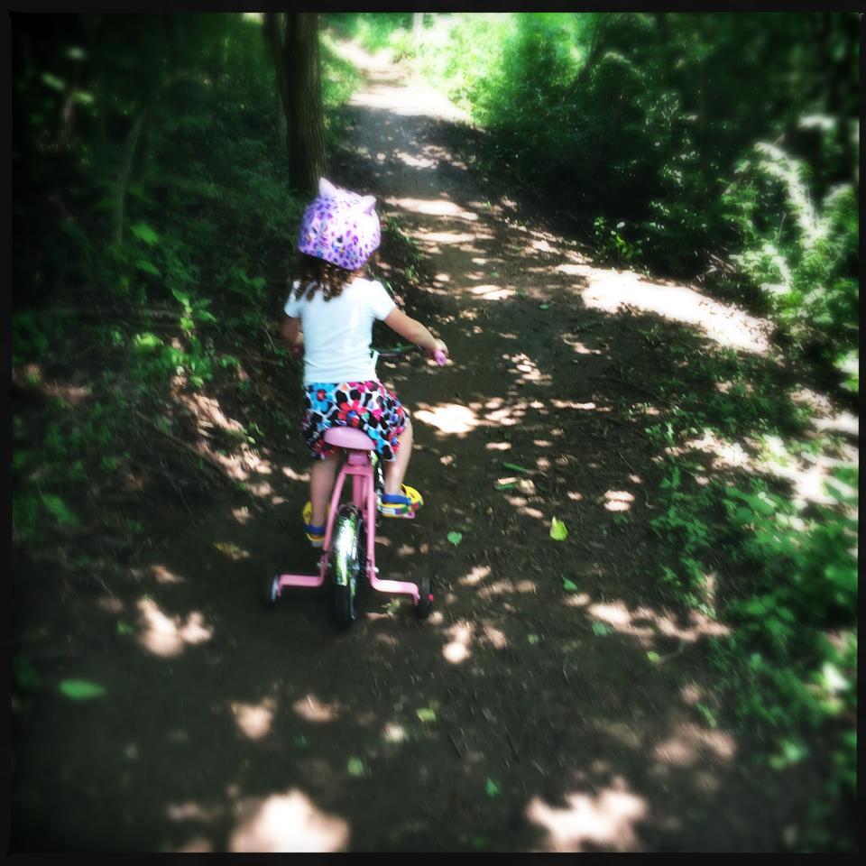 A young child rides a pink bicycle along a dirt trail surrounded by greenery. The child wears a white shirt, colorful patterned shorts, and a purple helmet with a playful design. Sunlight filters through the trees, creating a dappled effect on the path. Veterans Park mountain bike trail.