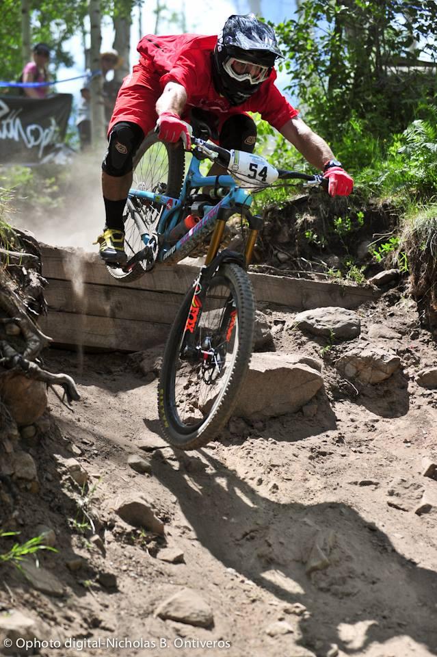 A mountain biker in a red jersey and helmet performs a jump off a wooden ramp, navigating a rocky dirt trail surrounded by greenery. Dust is kicked up behind the bike as it soars through the air. Bonzai DH mountain bike trail.
