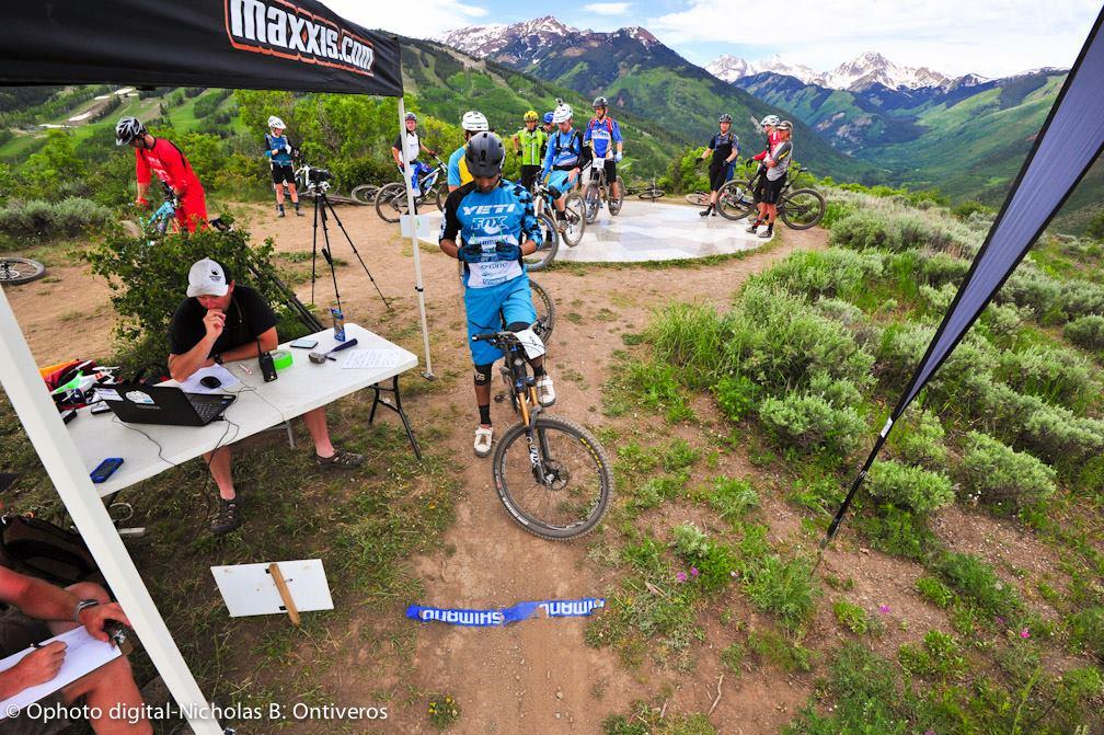 A group of mountain bikers in colorful jerseys gathers near a starting line in a scenic outdoor setting. One rider is preparing to start, while others observe. A person at a nearby table is using a laptop, possibly handling registrations or timing. The background features lush green mountains and a blue sky with scattered clouds, indicative of an event or competition taking place. Rim Trail mountain bike trail.