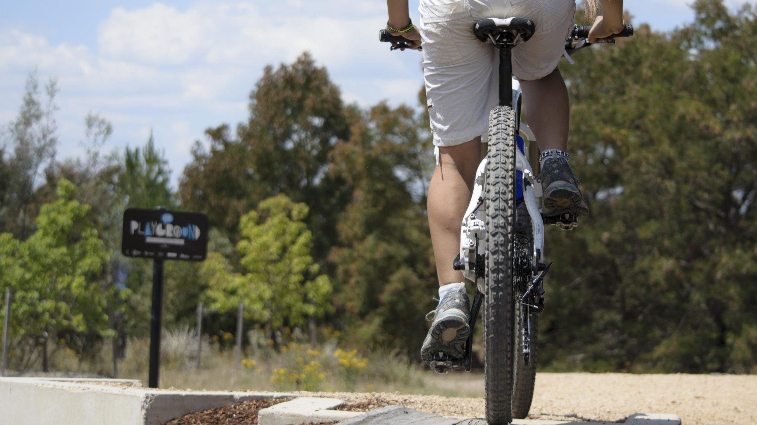 A person riding a mountain bike on a path near a playground sign, surrounded by trees and grassy areas under a blue sky. The focus is on the bike's rear wheel and the rider's legs. Stromlo Forest Park mountain bike trail.