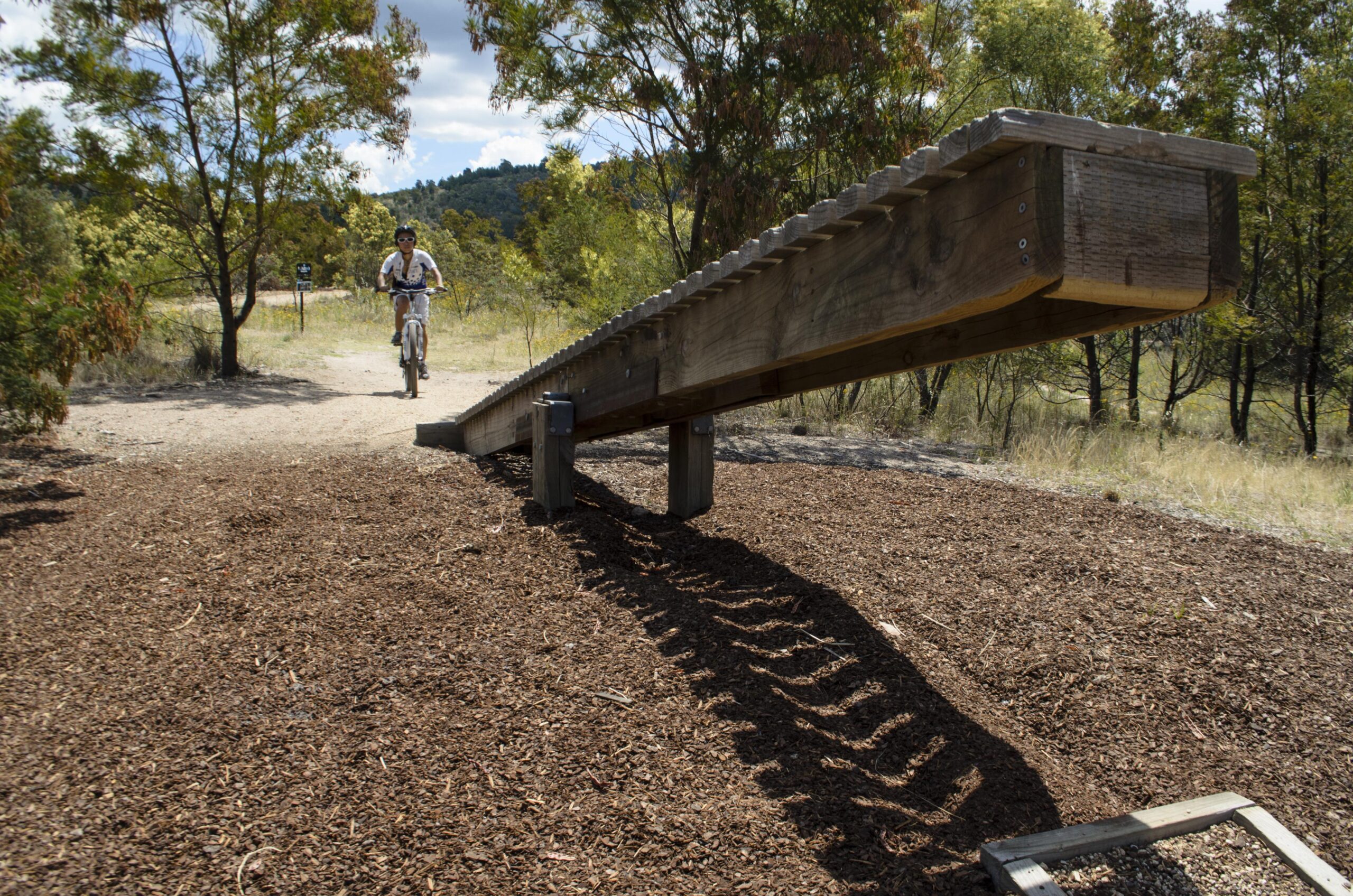 A person riding a bicycle approaches a wooden balance beam on a dirt path surrounded by trees and greenery. The wooden beam casts a shadow on the ground covered with wood chips, with a scenic backdrop of hills and a partly cloudy sky. Stromlo Forest Park mountain bike trail.