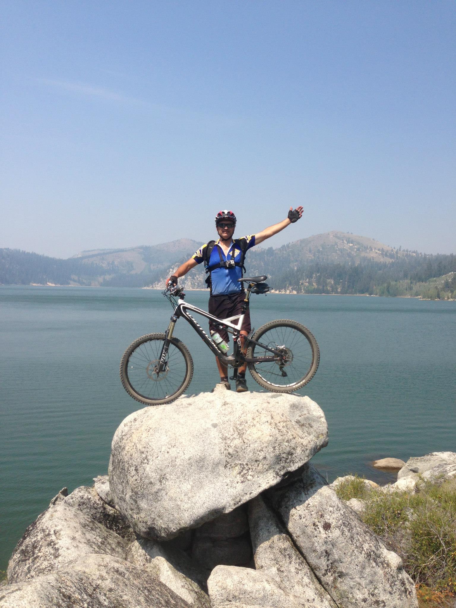A mountain biker stands on a large rock near the shore of a lake, with one arm raised in celebration. The biker is wearing a blue jersey, shorts, and a helmet, holding onto the bike beside him. In the background, the calm water of the lake reflects the surrounding mountainous landscape under a clear blue sky. Flume Trail mountain bike trail.