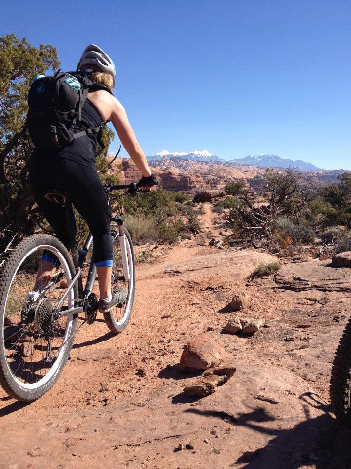 A cyclist riding a mountain bike on a rocky trail in a desert landscape, with mountains visible in the background under a clear blue sky. Captain Ahab mountain bike trail.