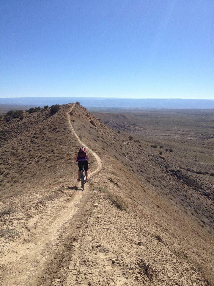 A cyclist riding along a narrow dirt trail on a hillside, surrounded by a vast, open landscape under a clear blue sky. The path winds along the ridge, with rolling hills and sparse vegetation visible in the distance. 18 Road Trails / North Fruita Desert mountain bike trail.
