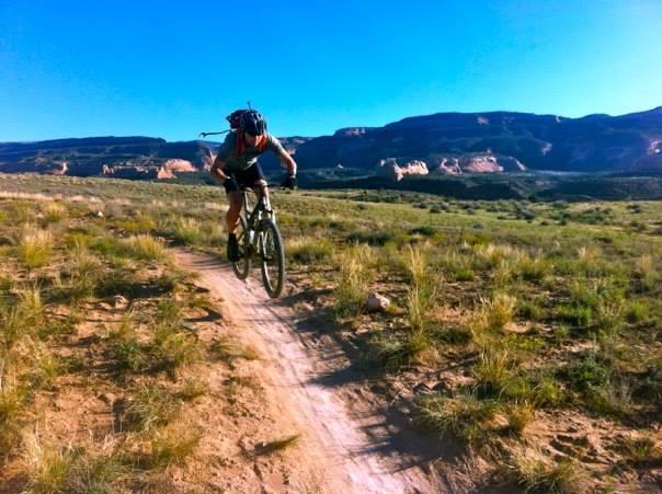 A mountain biker jumping on a narrow dirt trail in a grassy landscape, with rocky hills and a clear blue sky in the background. Mary's Loop / Horsethief Bench mountain bike trail.