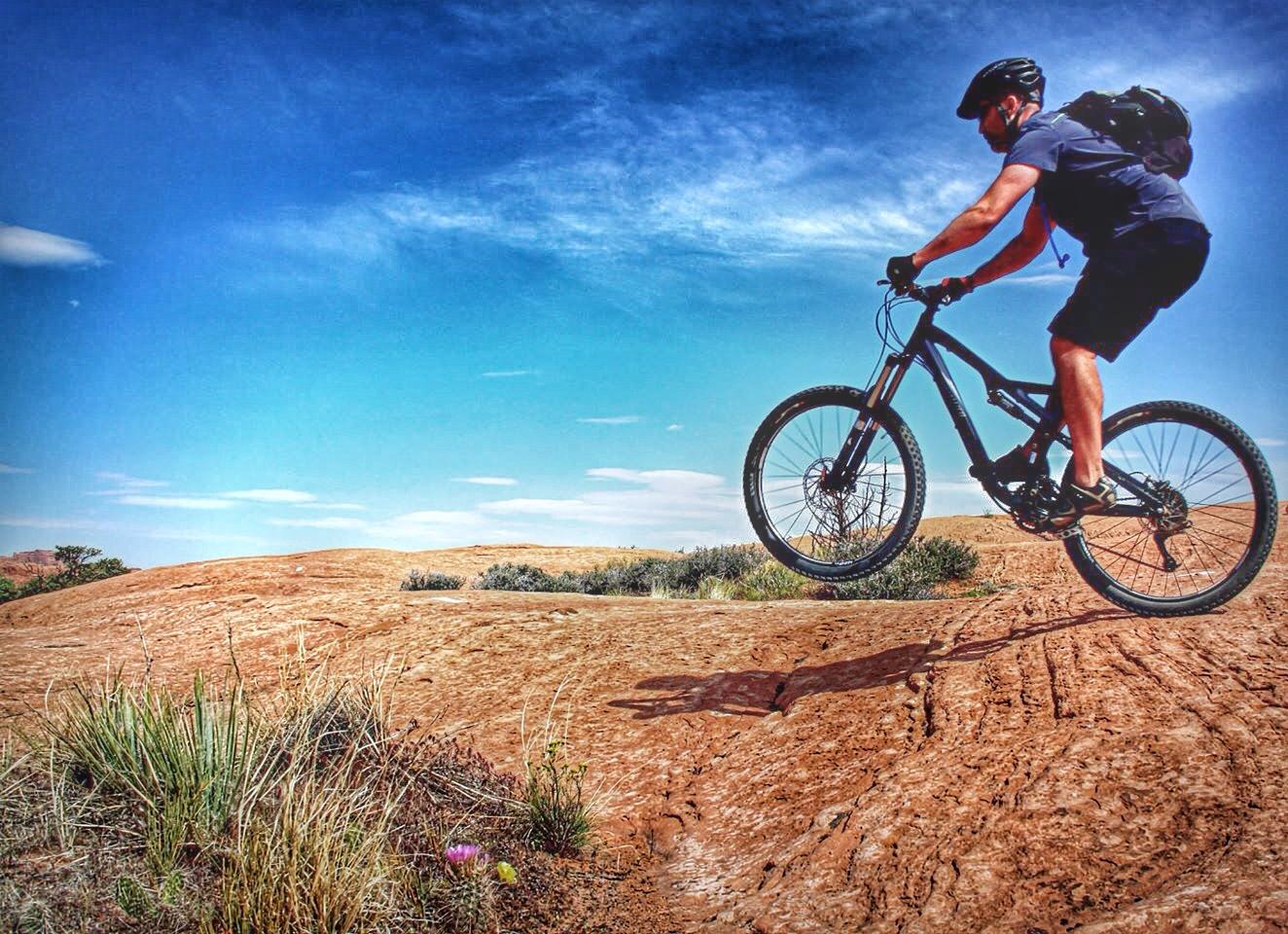A mountain biker performing a jump over rocky terrain under a clear blue sky. The landscape features reddish-brown earth and sparse vegetation, with a few patches of grass and cacti visible in the foreground. Slickrock mountain bike trail.