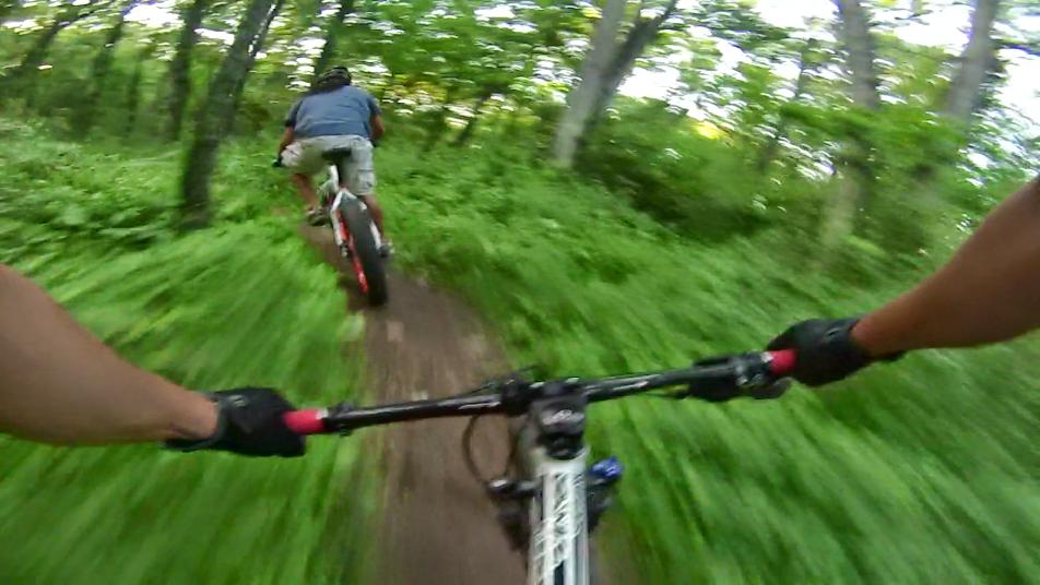 Two mountain bikers riding through a wooded trail, with one cyclist in the foreground holding the handlebar and the other ahead, slightly out of focus. The scene is vibrant with lush green foliage surrounding the path, creating a sense of speed and adventure. Raceway Woods mountain bike trail.