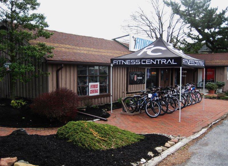 Front view of a store called "Fitness Central," featuring a tent with the same name and a variety of bicycles displayed outside. The building has a rustic design with a brick pathway and landscaped greenery.