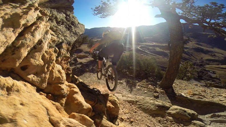 A mountain biker navigating a rocky trail with sunlight shining in the background, surrounded by trees and mountainous terrain. Red Rocks / Dakota Ridge mountain bike trail.