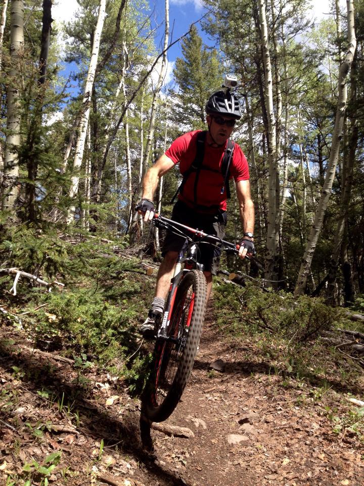 A mountain biker riding on a forest trail, balancing on one wheel. The cyclist wears a helmet and a red shirt, surrounded by trees and foliage under a blue sky with scattered clouds. Rainbow Trail: Silver Creek to Hwy 285 mountain bike trail.
