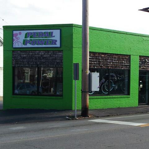 A bright green storefront with the sign "Pedal Power" in purple letters. Large windows display bicycles and accessories. A utility pole stands beside the building, and the scene is set on a street with vehicles visible in the background.