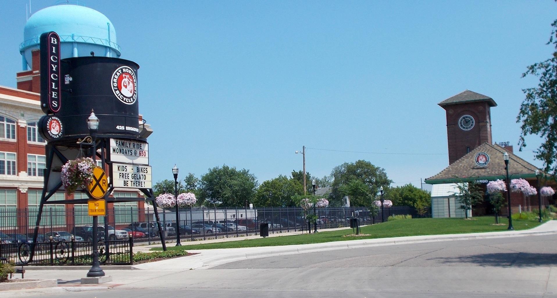 A sunny street scene featuring a large bicycle-themed water tower sign and a brick clock tower in the background. The foreground includes street signs and blooming flower baskets, along with a parked bicycle. In the background, a community event advertisement is displayed on the water tower, highlighting family bike rides and free gelato for kids.