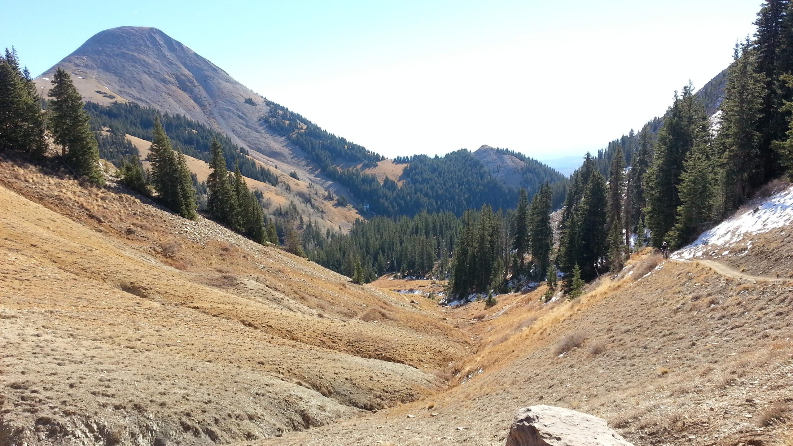 A scenic view of a mountainous landscape featuring a prominent peak in the background, surrounded by sloping hills and patches of forest. The foreground shows dry, grassy terrain with scattered rocks, leading into a valley lined with evergreen trees. The sky is clear, indicating a bright and sunny day. The Whole Enchilada mountain bike trail.