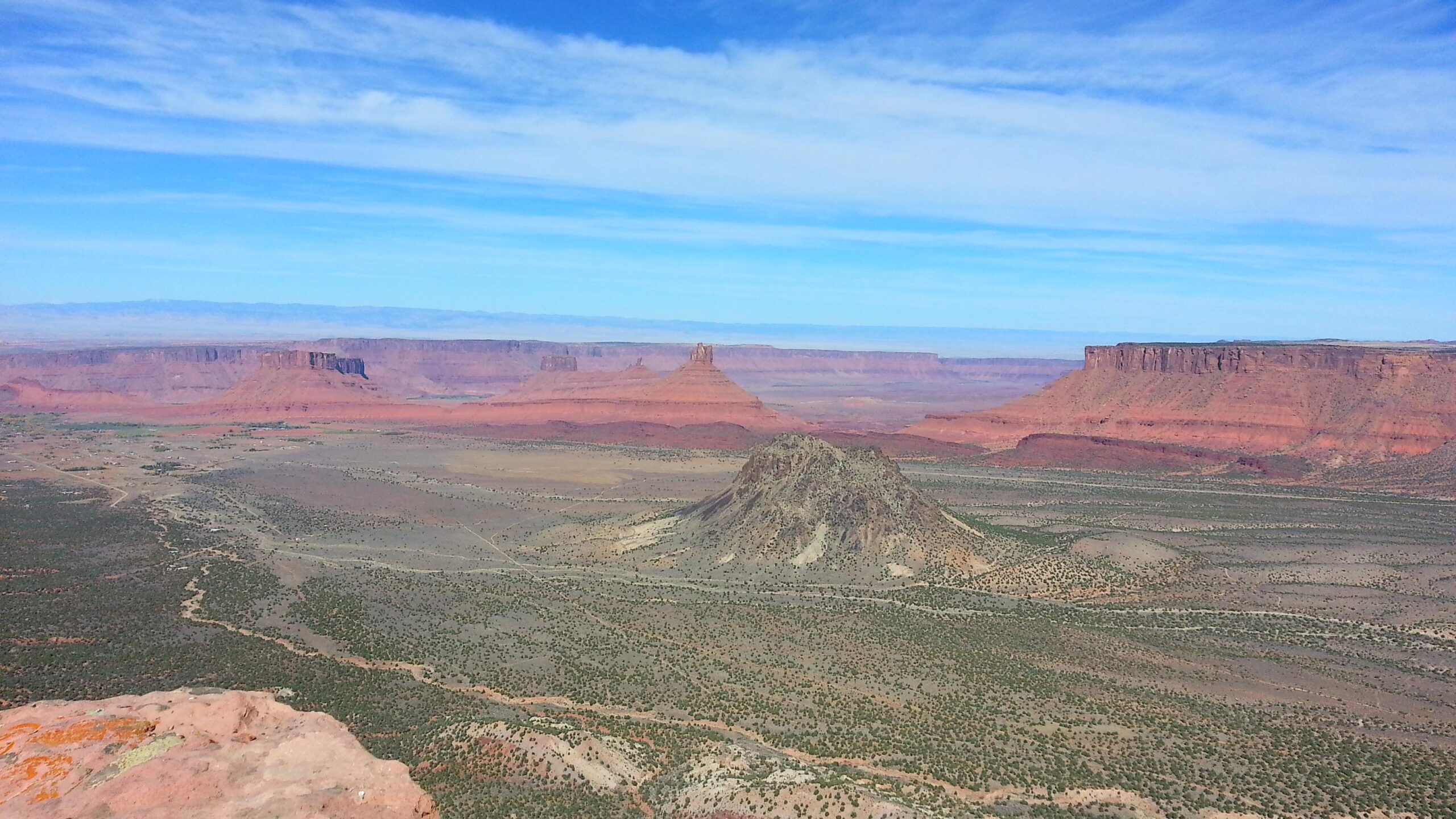 A panoramic view of a desert landscape featuring red rock formations and distant mesas under a blue sky with scattered clouds. The foreground includes a rocky outcrop with sparse vegetation, while the background showcases layered cliffs and mesas in varying shades of red and brown. The Whole Enchilada mountain bike trail.