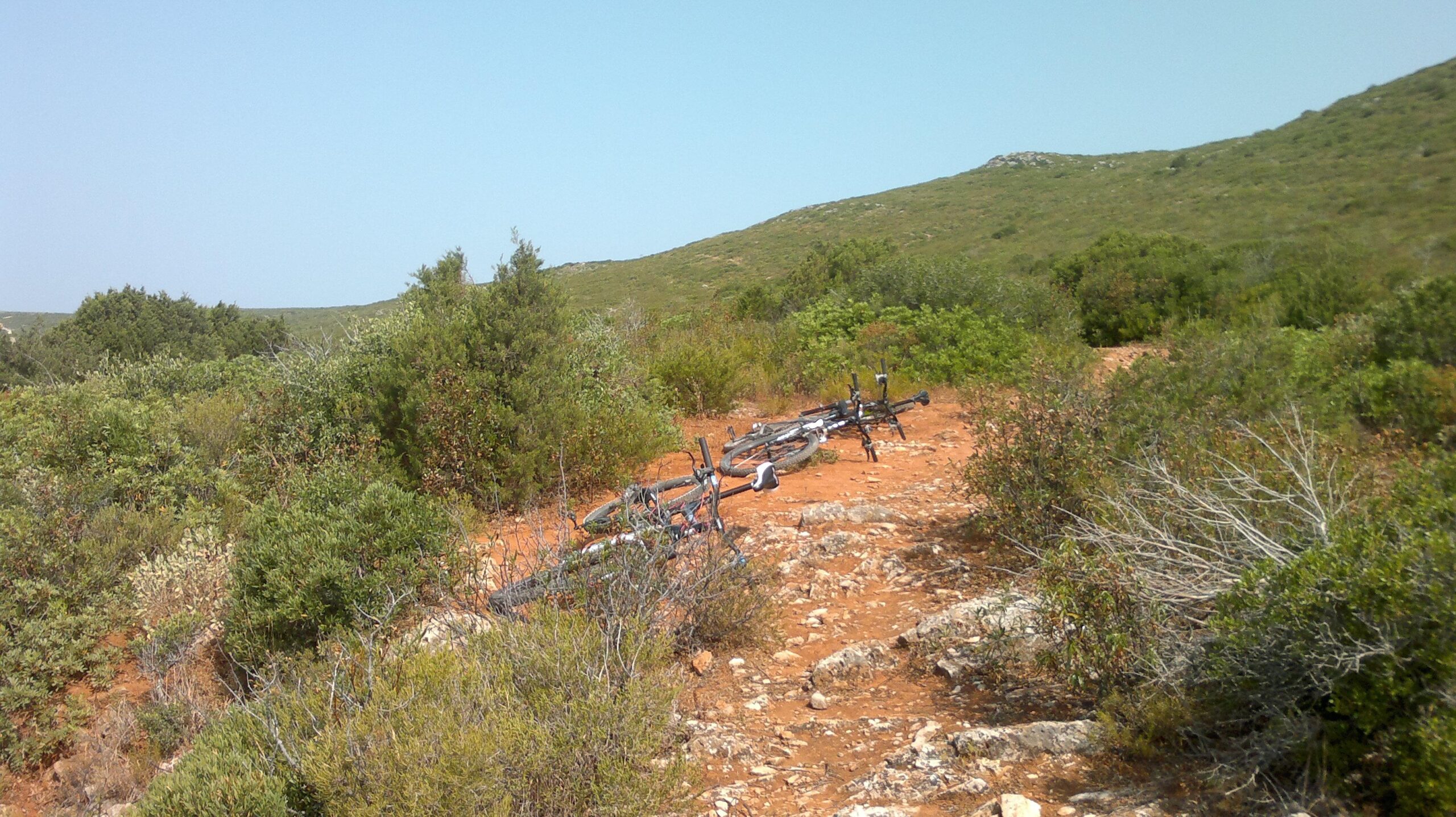 A dirt path surrounded by greenery, featuring a few mountain bikes lying on the ground. The landscape includes rolling hills and sparse vegetation, with a clear blue sky above. Arrabida mountain bike trail.