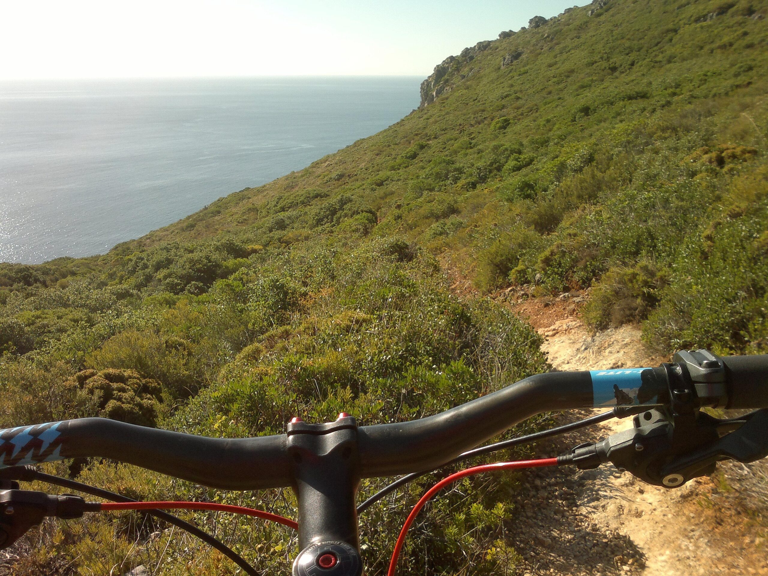 Mountain bike handlebars in the foreground with a scenic view of a coastal landscape, including greenery and the ocean in the background. The trail ahead winds down the hillside, framed by lush vegetation and rocky cliffs. Arrabida mountain bike trail.