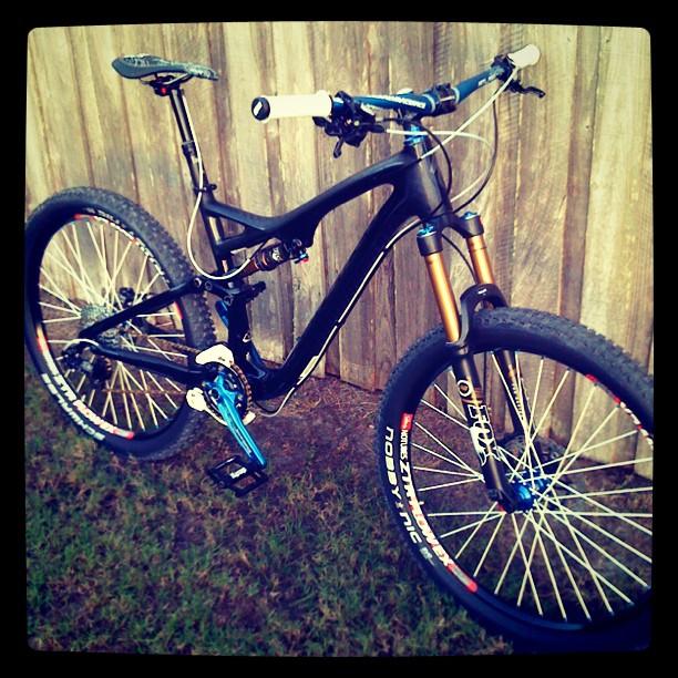 Specialized Stumpjumper FSR Carbon EVO: A black mountain bike with blue and orange accents, featuring a prominent suspension fork and wide tires, positioned on grass with a wooden fence in the background. The bike has white handlebars and is equipped with a rear shock absorber.