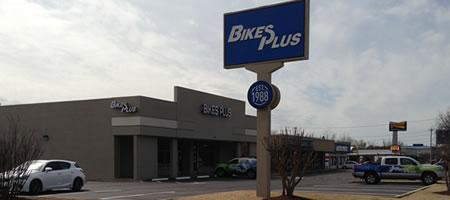 A storefront for "Bikes Plus," featuring a large blue sign with the business name. The building is a modern, single-story structure, with parked vehicles in front. The sky is partly cloudy, suggesting a daytime scene.