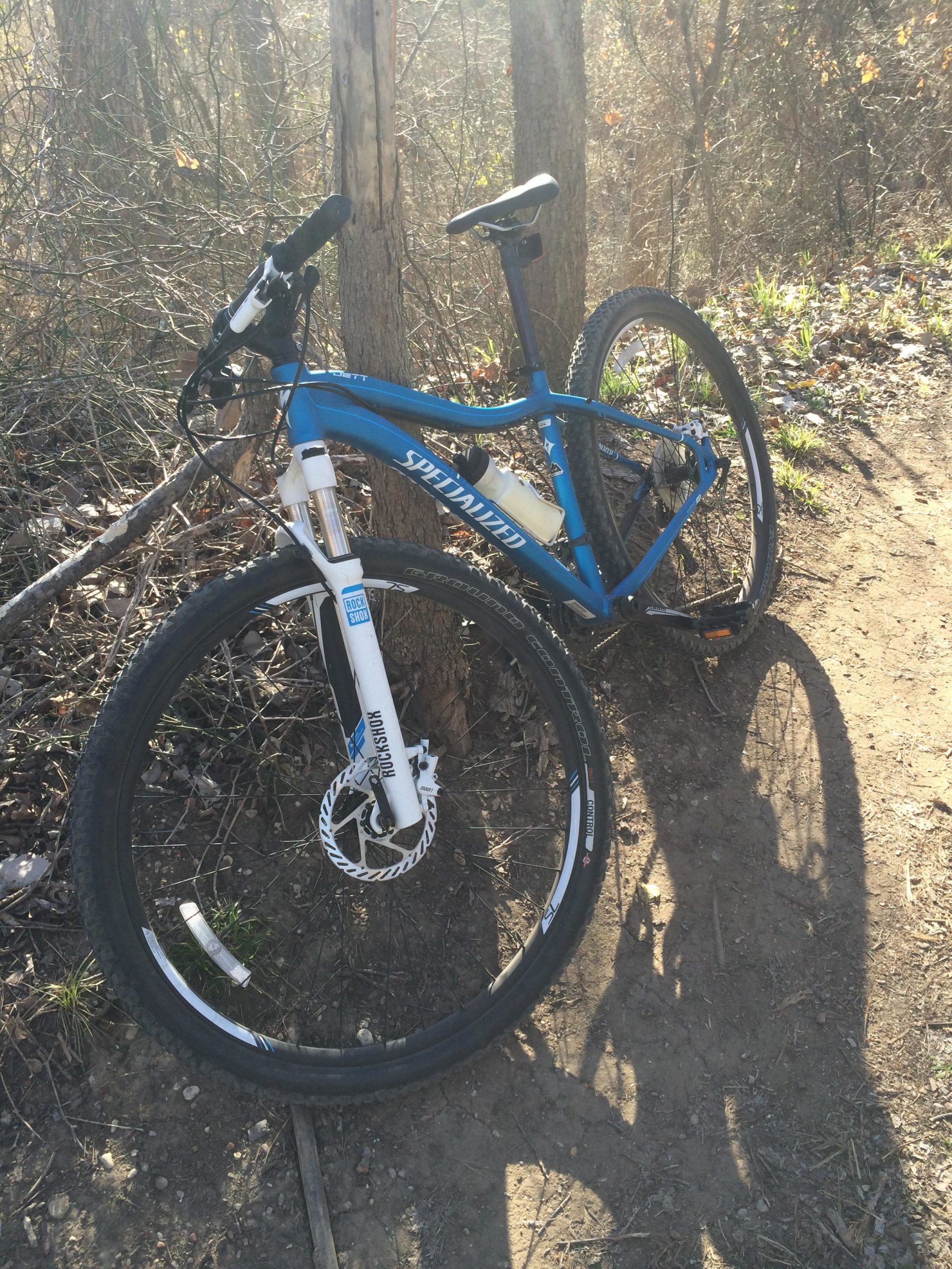 A blue mountain bike leaning against a tree, positioned on a dirt trail surrounded by greenery and sunlight filtering through the trees. River Legacy mountain bike trail.