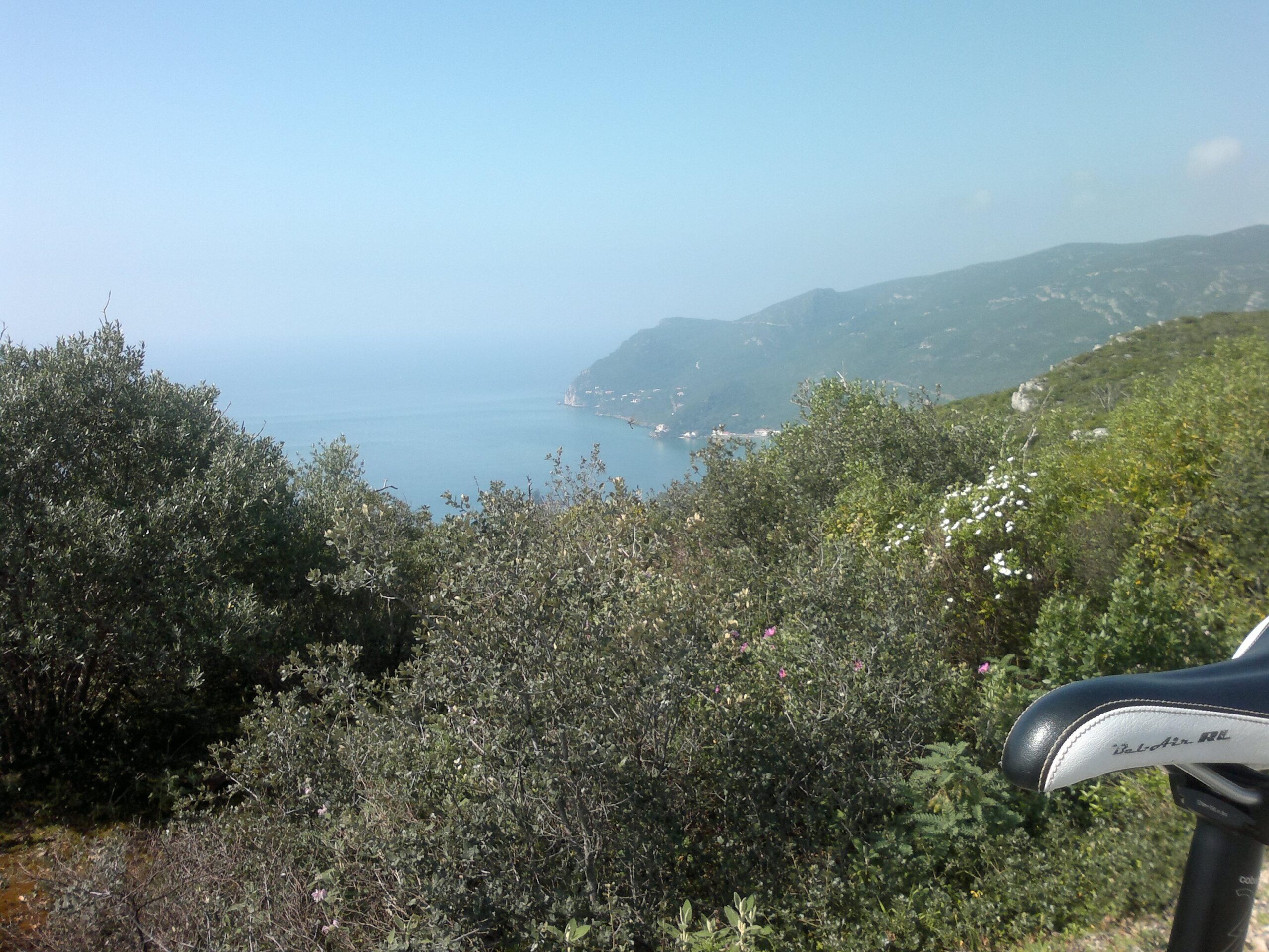 A scenic view from a mountainous area overlooking a calm sea, with greenery in the foreground and rugged hills in the background. A bicycle seat is partially visible on the right side of the image. The sky is clear and blue, creating a bright and inviting atmosphere. Arrabida mountain bike trail.