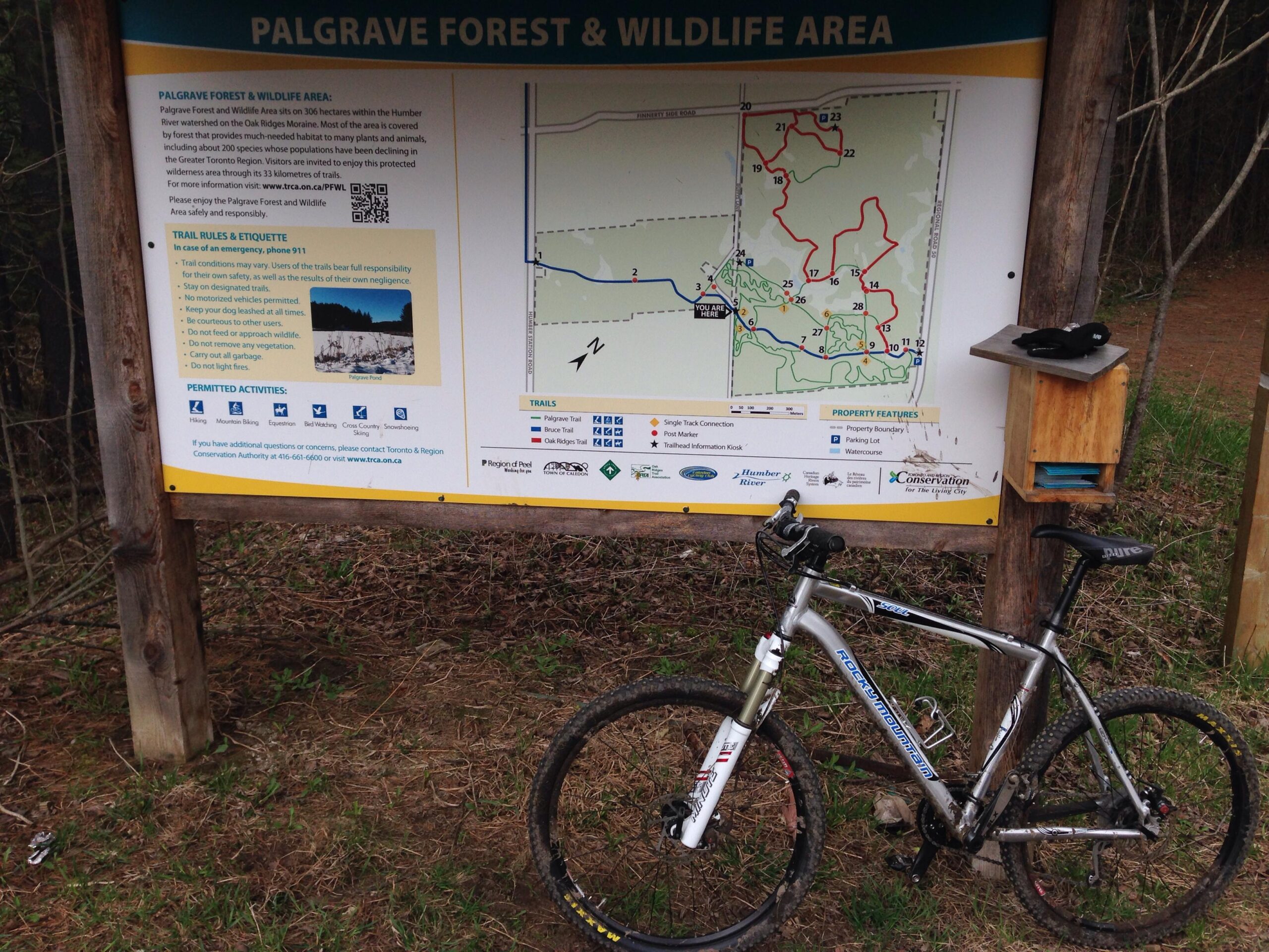 A mountain bike parked next to a large informational sign detailing the Palgrave Forest and Wildlife Area. The sign features a map of the trails, rules, and permitted activities such as hiking, mountain biking, and cross-country skiing. The surrounding area is wooded with grass and dirt paths visible. Palgrave Trail mountain bike trail.