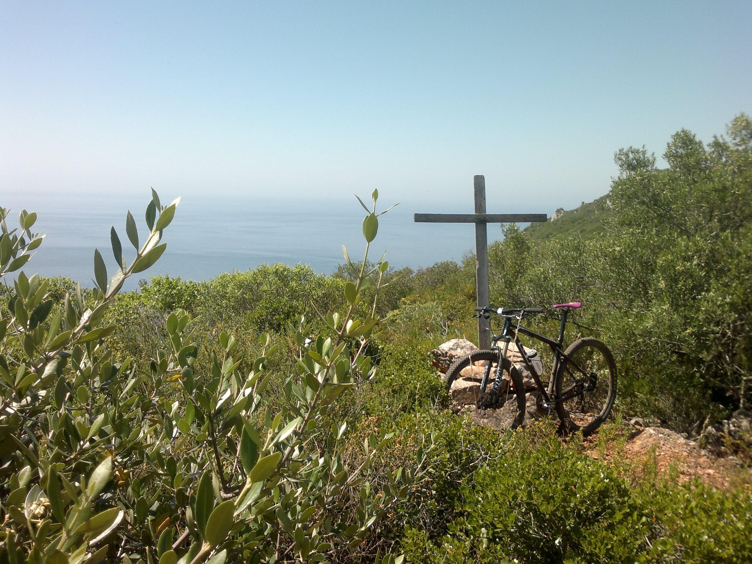 A mountain bike resting beside a wooden cross, surrounded by lush greenery and overlooking a calm ocean under a clear blue sky. Arrabida mountain bike trail.